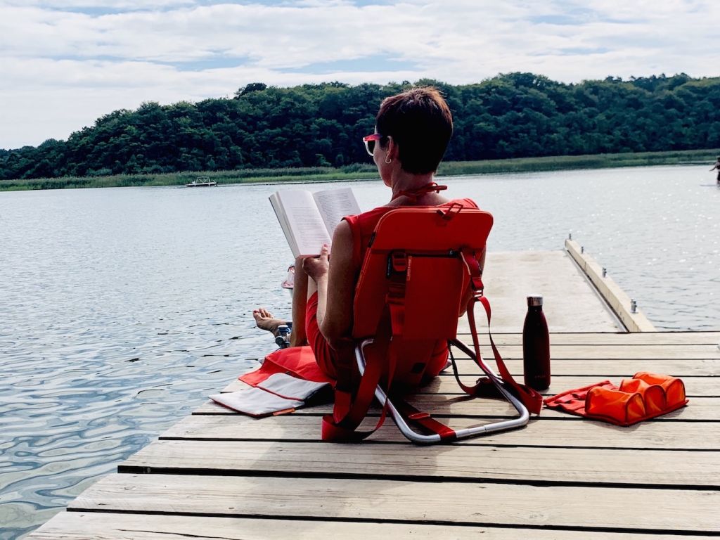 Frau entspannt Buch lesend mit Rückenlehne am See