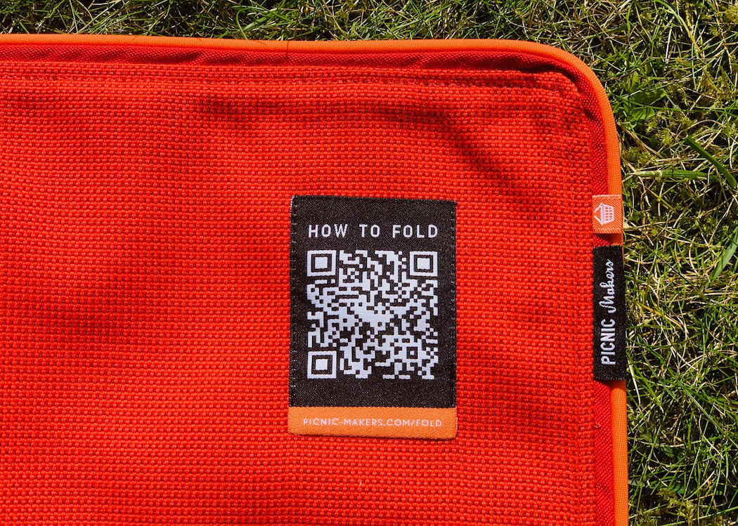 Close-up of the “How to Fold” woven label, with side-sewn flag labels, a white picnic basket on an orange background, and a black flag label reading “picnic.makers” in white.